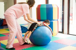 Female therapist pushing a boy on top of a stability ball to help him relax in a therapy center